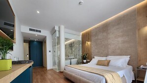 Una habitación de hotel moderna con una cama grande, suelo de madera, cabina de ducha de vidrio y una pequeña zona de escritorio.