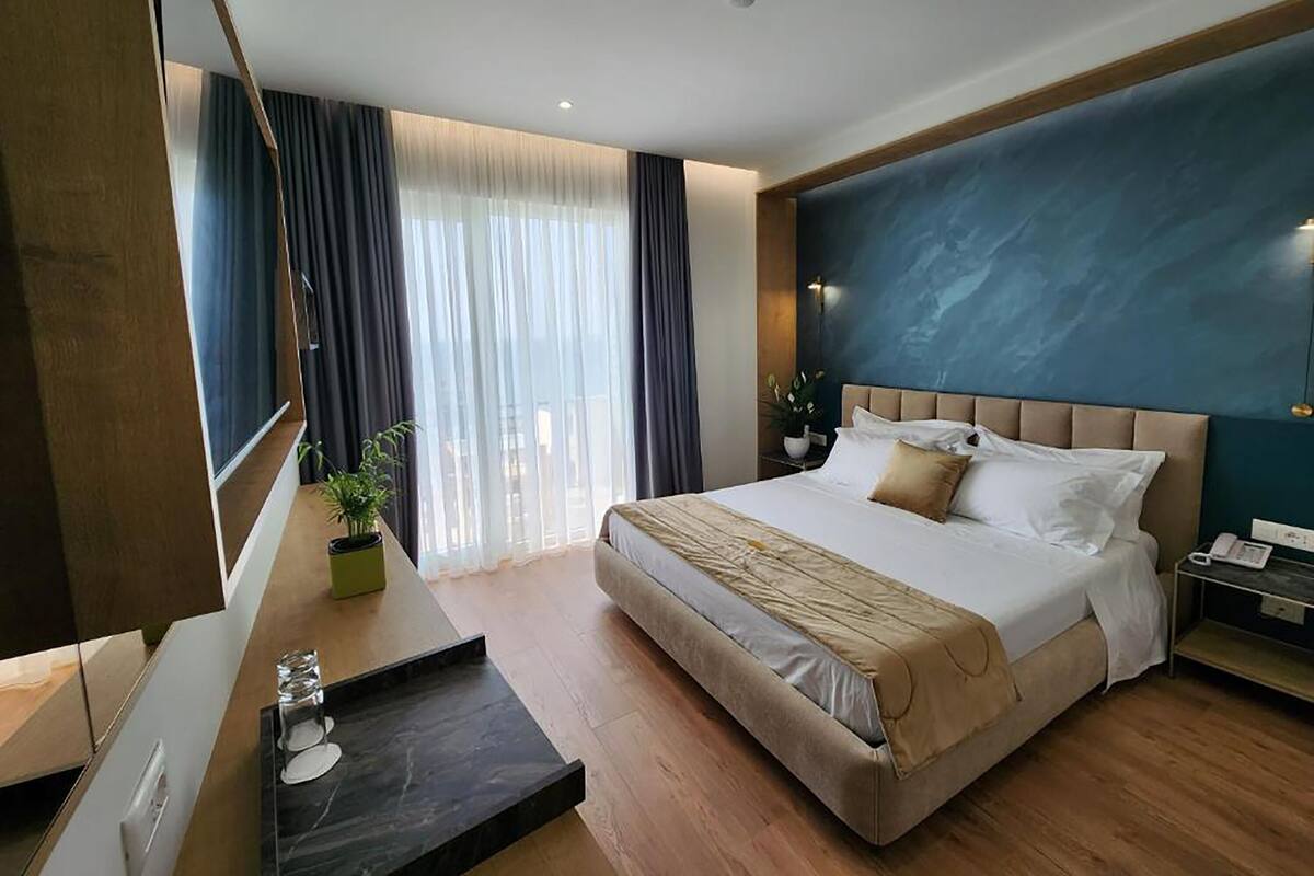 A modern hotel room with a large bed, a bedside table, a mirror, and a view of the outdoors through large windows.