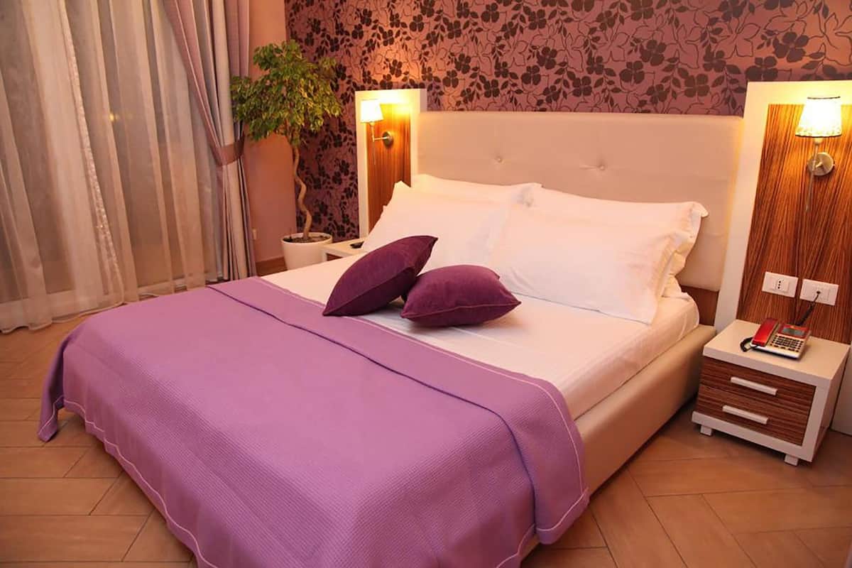 A neatly made bed with a purple bedspread and two pillows, a bedside lamp, a nightstand with a phone, and a wall-mounted lamp.