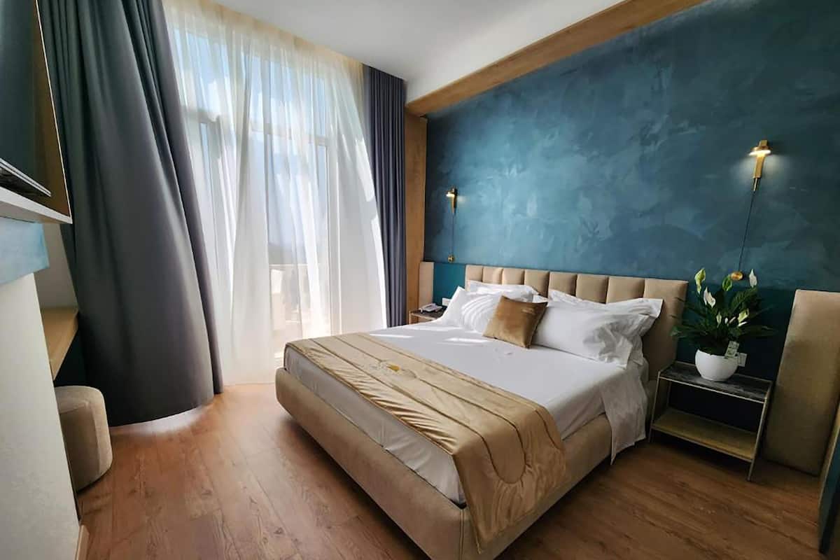 A modern hotel room with a large bed, a bedside table with a lamp, and a view of the outdoors.