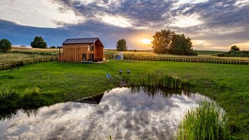 Un granero de madera rústico con techo de metal, un pequeño estanque que refleja el cielo y un campo cubierto de hierba con una cerca de madera.