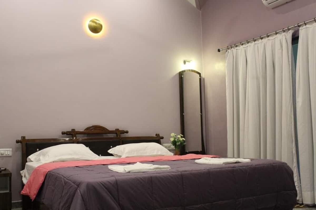 Una cama bien hecha con una colcha morada y almohadas blancas, una mesita de noche con una lámpara y una ventana con cortinas blancas.