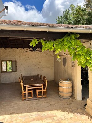 A rustic outdoor patio with a wooden table and chairs, a barrel, and a pergola with green vines.