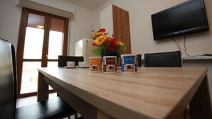 A hotel room with a wooden dining table, three coffee containers, a vase with flowers, a flat-screen TV mounted on the wall, and a window with blinds. - Renovated Flat Near the Beach - Beahost (Rosolina)