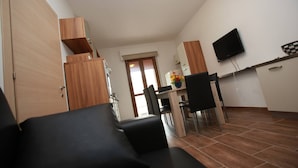A modern living room with a black sofa, wooden cabinets, a dining area with a table and chairs, and a flat-screen TV mounted on the wall. - Renovated Flat Near the Beach - Beahost (Rosolina)