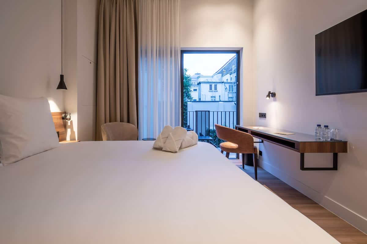 Ein modernes Hotelzimmer mit einem großen Bett, einem Schreibtisch, einem Sessel, einem an der Wand befestigten Fernseher und einem Balkon mit Blick auf Gebäude.