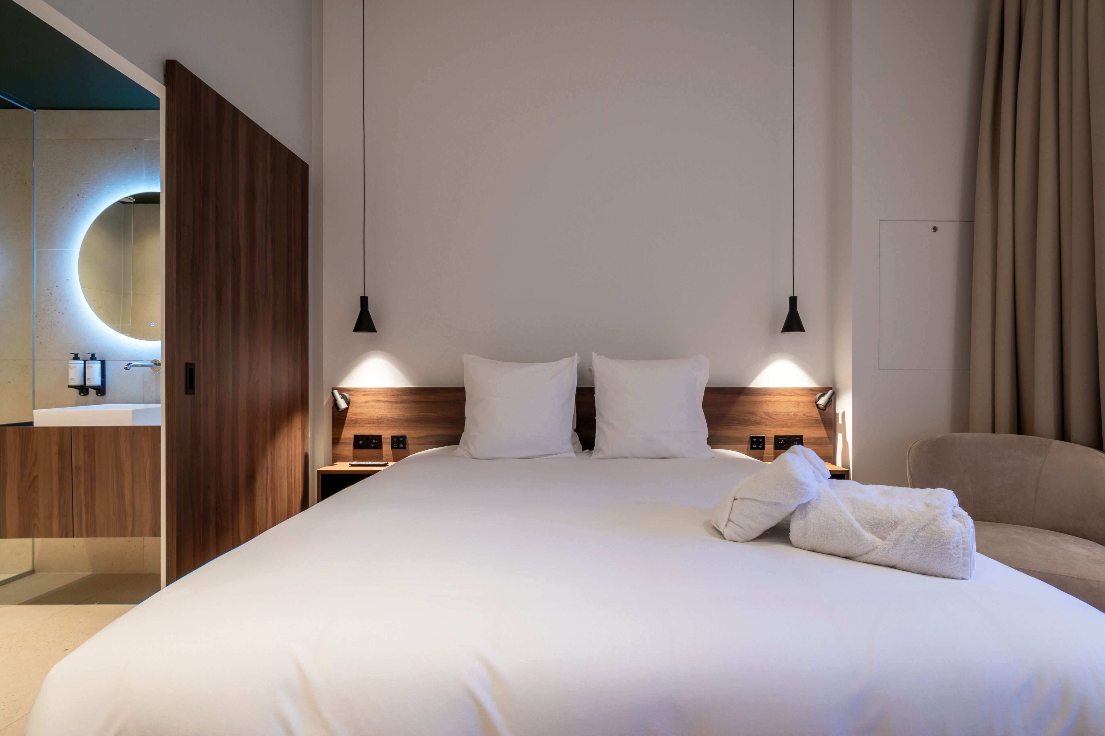 Een moderne hotelkamer met een groot bed, twee nachtkastjes, een houten hoofdbord en een ronde spiegel boven het bed.
