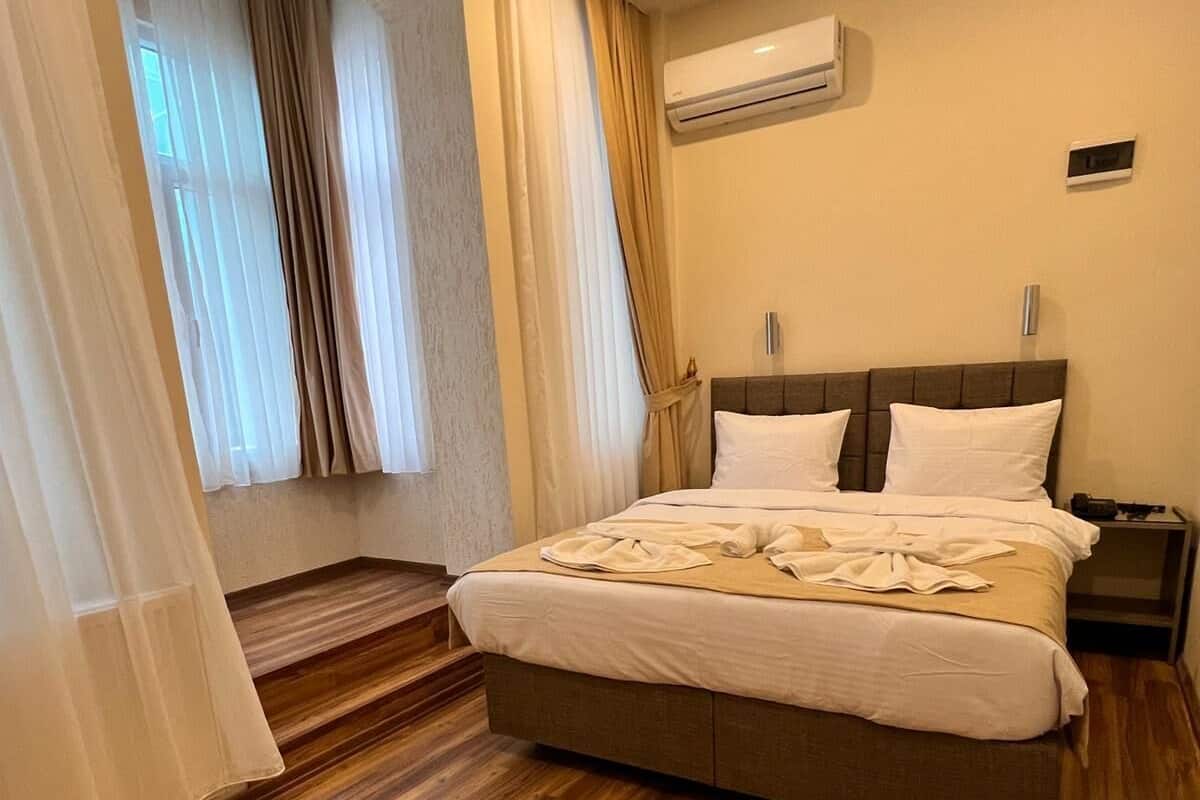 A hotel room with a large bed, wooden flooring, an air conditioning unit, and a window with curtains.
