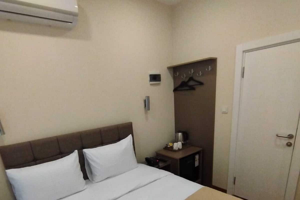 A hotel room with a bed, a bedside table, an air conditioning unit, a coat rack, and a door.