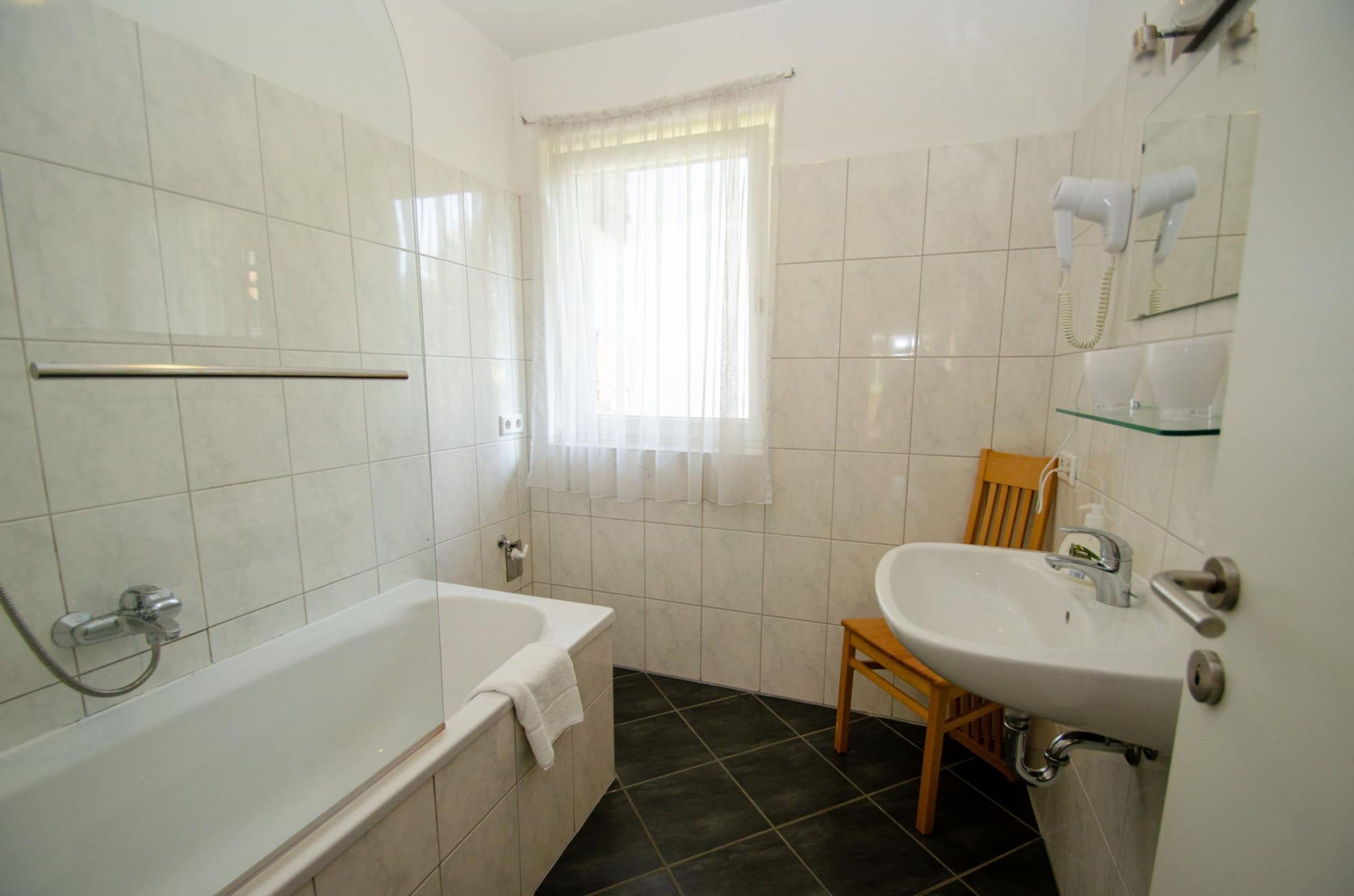 Een badkamer met een bad, een wastafel, een stoel, een raam met gordijnen en een haardroger aan de muur.