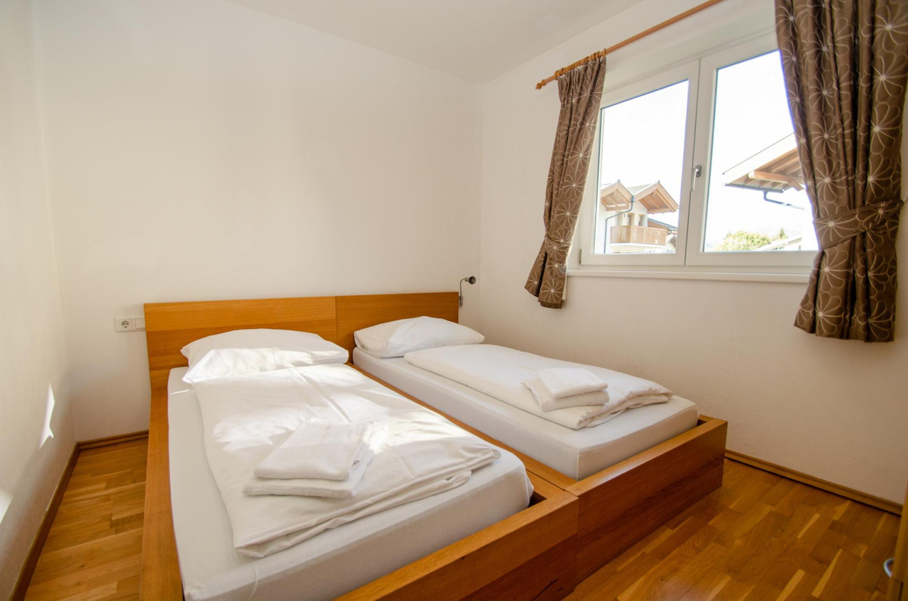 Un dormitorio con dos camas individuales, cabeceras de madera, una ventana con cortinas y vistas a casas.