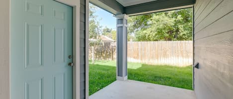 Un porche abrité avec une porte bleu clair, un sol en béton et une vue sur une cour arrière agrémentée d’une clôture en bois et d’une pelouse.
