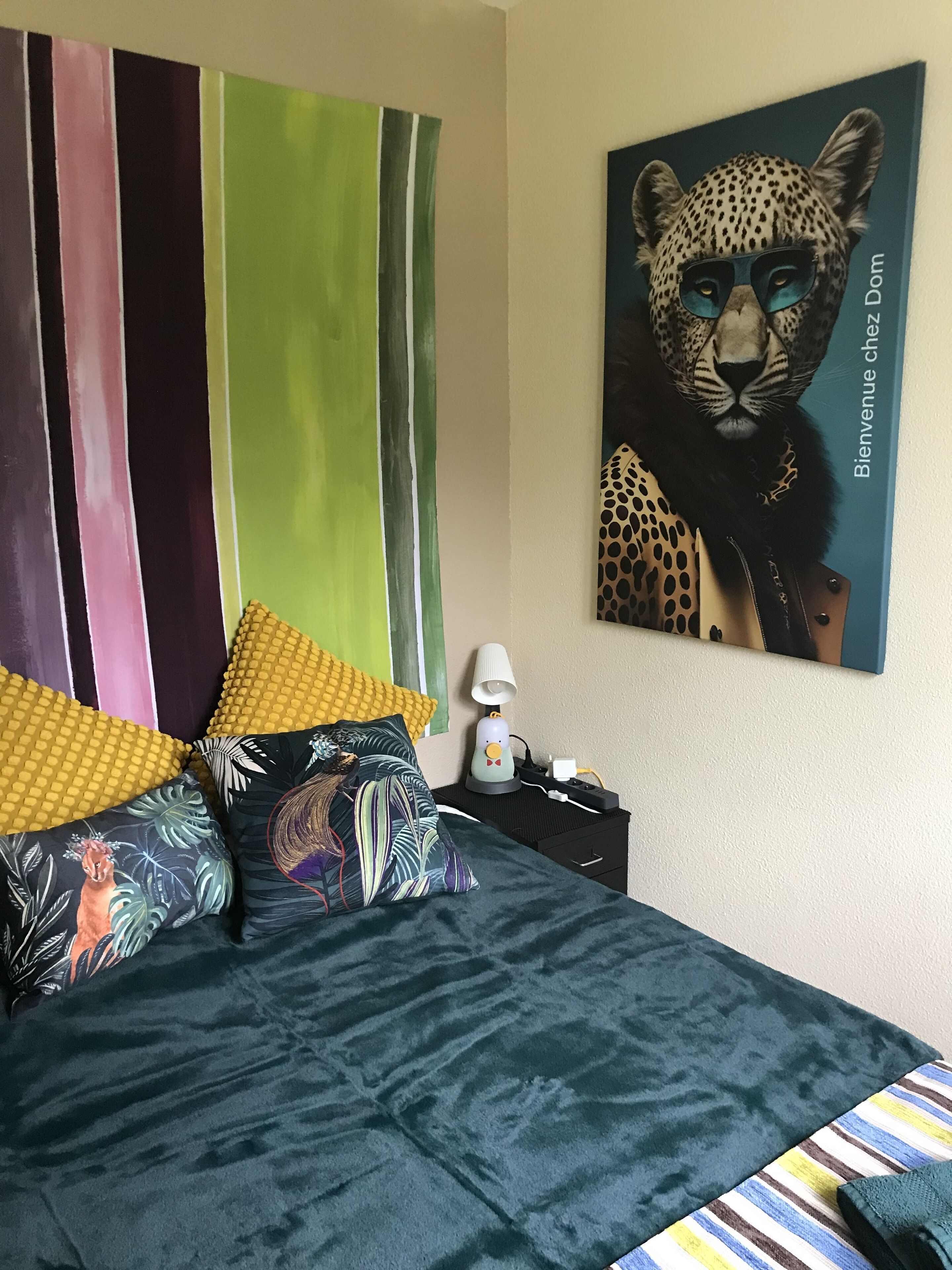Una cama con un edredón verde oscuro y almohadones coloridos, una lámpara en una mesita de noche y un cuadro con motivos de leopardo en la pared.