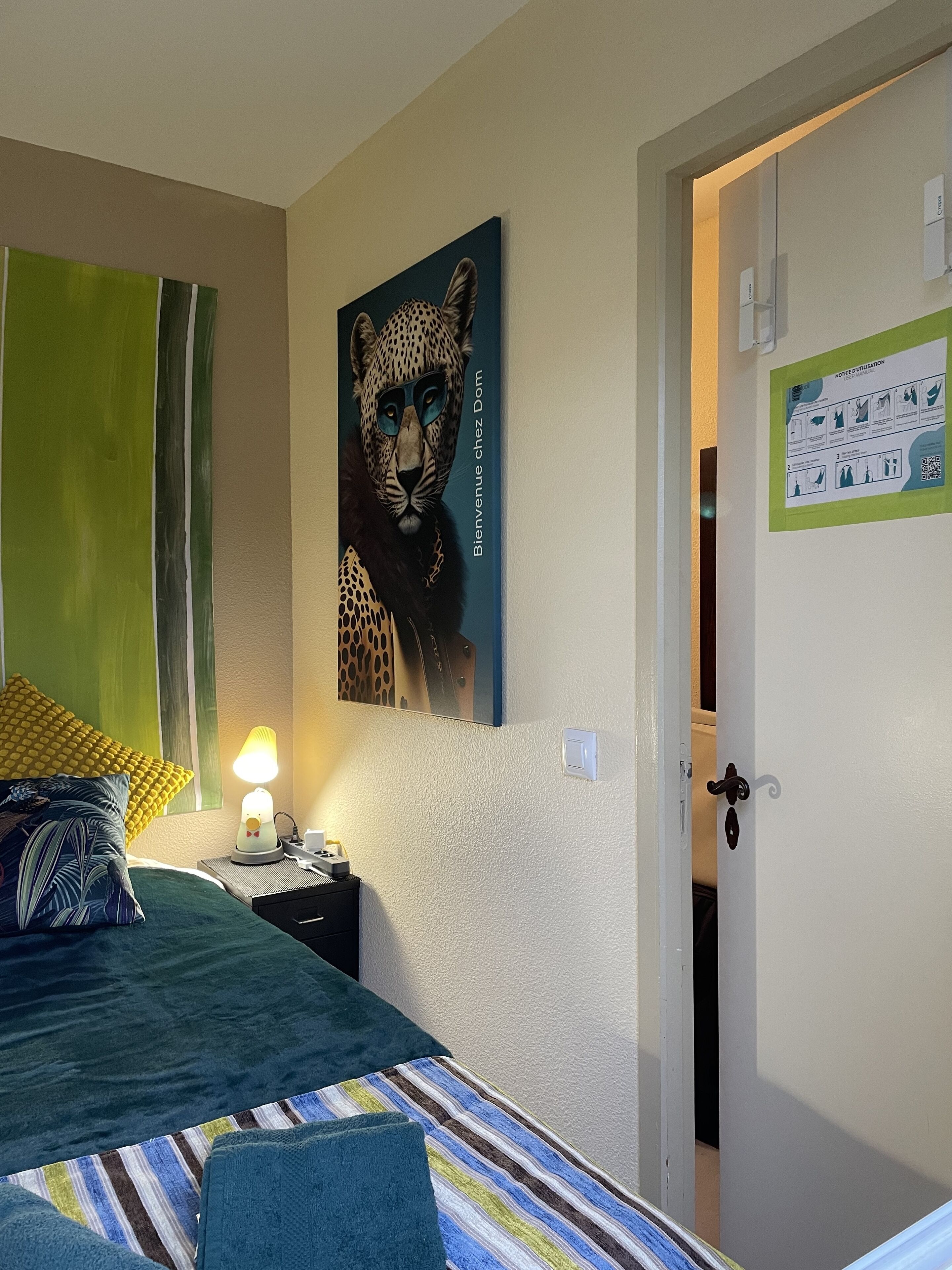A bedroom with a bed, a nightstand, a lamp, a leopard artwork, a green curtain, and a door with a notice.