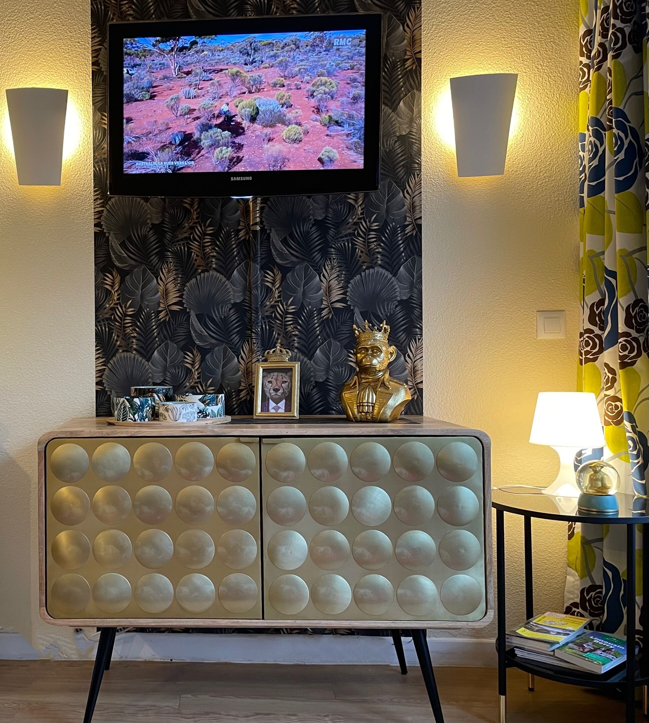 Una sala de estar con una televisión de pantalla plana montada en la pared, un mueble decorativo con una imagen de un león y una mesita con una lámpara y libros.