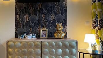 Una sala de estar con una televisión de pantalla plana montada en la pared, un mueble decorativo con una imagen de un león y una mesita con una lámpara y libros.