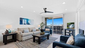A living room with a sofa, coffee table, and a TV on a stand. There is a ceiling fan and a window with a view of the city. - Harbor Cottage North Coast Village G36-Ocean Views-*Super Close To Beach* (Oceanside)