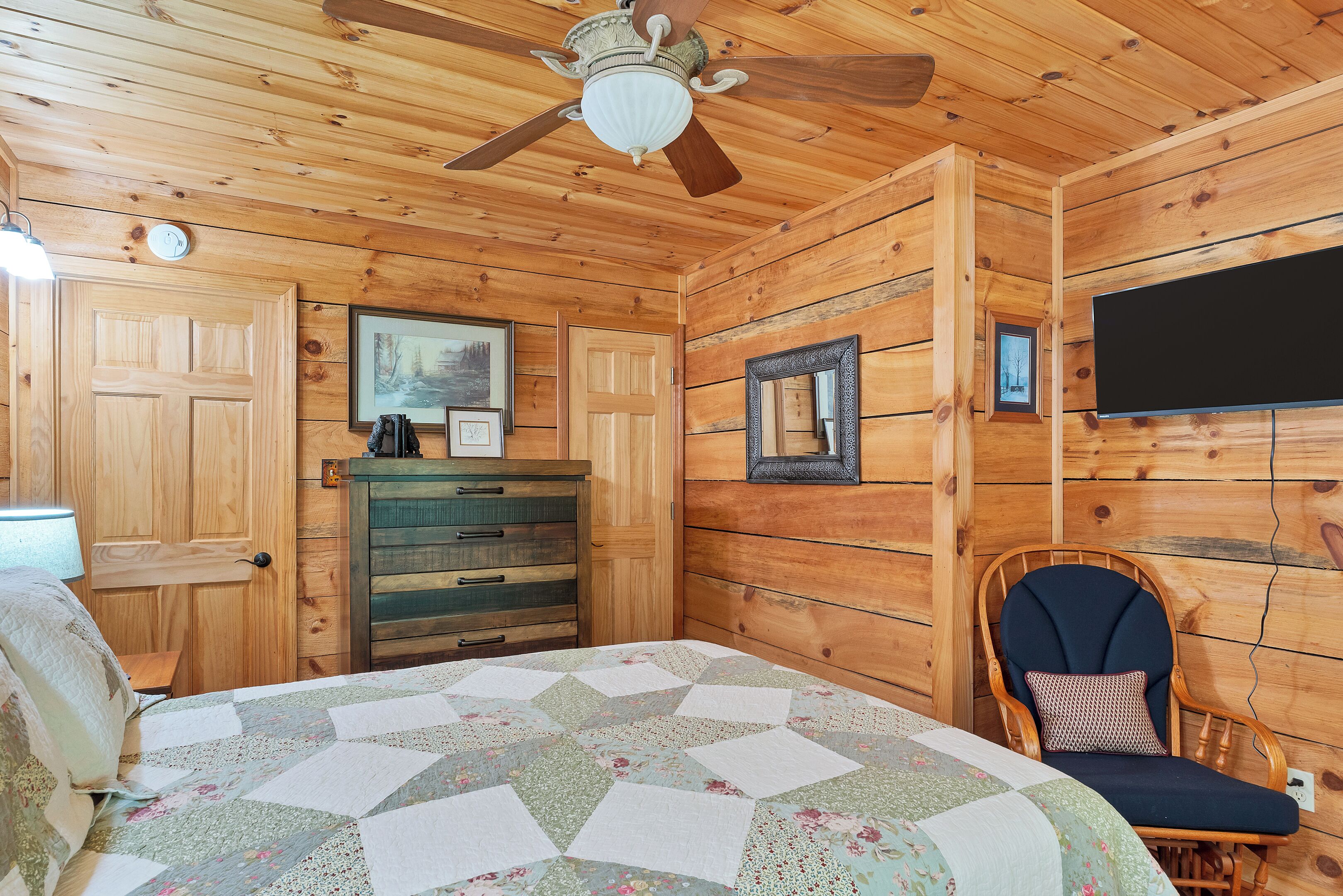 A wooden cabin bedroom with a bed, dresser, chair, ceiling fan, and a TV mounted on the wall.