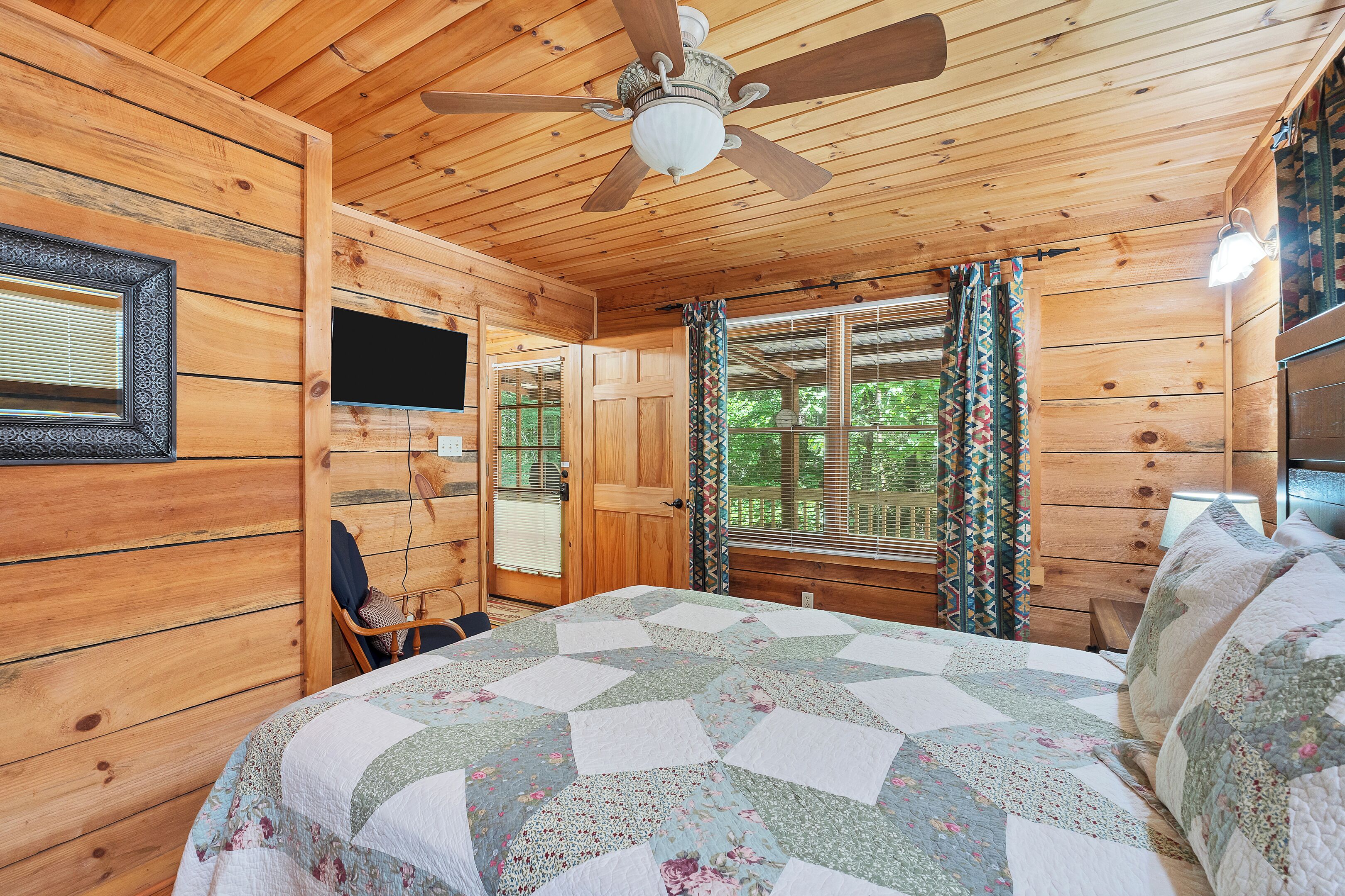 Une chambre dans un chalet en bois, avec un lit, un ventilateur de plafond, une télévision et une fenêtre avec des rideaux.
