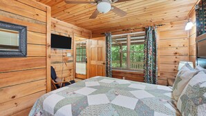 A wooden cabin bedroom with a bed, a ceiling fan, a TV, and a window with curtains. - SECLUDED♤Bonfire pit♤2 bdrm/2bath♤Pet friendly (Townsend)
