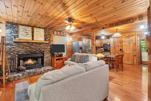 A cozy cabin interior with a stone fireplace, wooden ceiling, and a dining area.