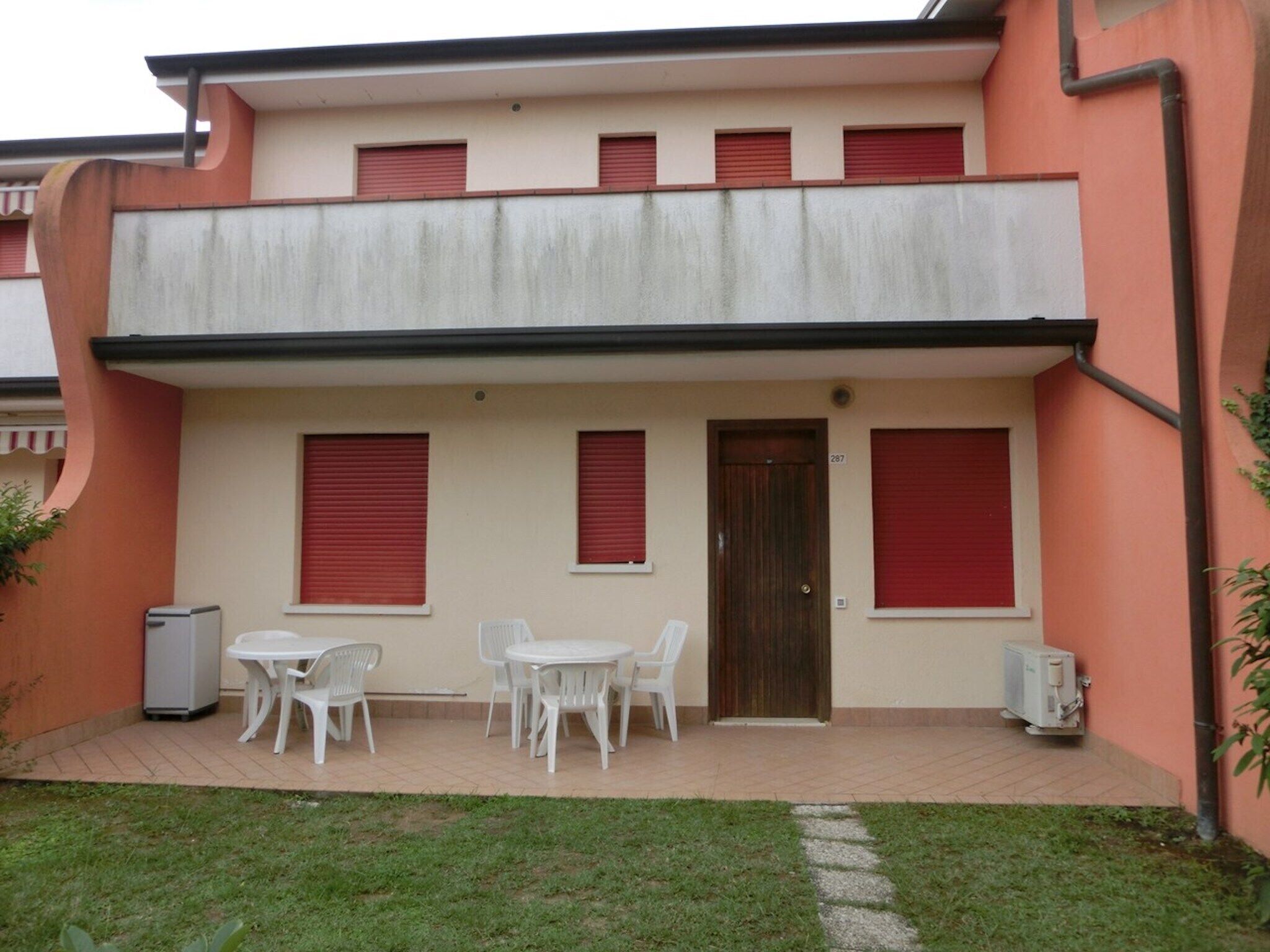 Un edificio de dos pisos con techo plano, una puerta principal y un pequeño patio con sillas y una mesa de plástico blanco.