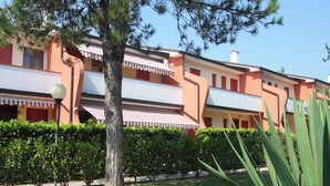 A building with a red and white facade, multiple balconies, and a well-maintained garden with a tree and shrubs. - Functional Flat in a Fantastic Residence With Pool (San Michele al Tagliamento)