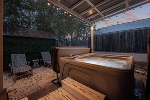 A hot tub with a wooden deck, surrounded by wicker chairs and a small table, under a covered area with string lights. - Private Wellness retreat|Hottub|Sauna|Prime loc (Fredericksburg)