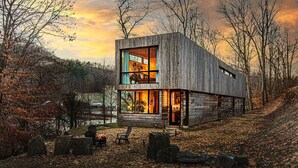 A modern wooden cabin with a large glass window, a fire pit, and a view of the surrounding forest. - Architect designed w/ Sauna, Hot Tub, Fire Pit, Swings, Game Rm,3 near Asheville (Mars Hill)