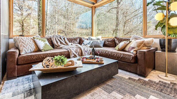 A cozy living room with a brown leather sofa, a coffee table with a fruit platter, and a large window offering a view of trees. - Architect designed w/ Sauna, Hot Tub, Fire Pit, Swings, Game Rm,3 near Asheville (Mars Hill)