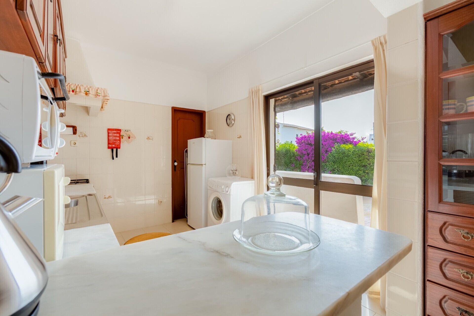 A compact kitchen with a washing machine, a marble countertop, and a view of a garden through a window.