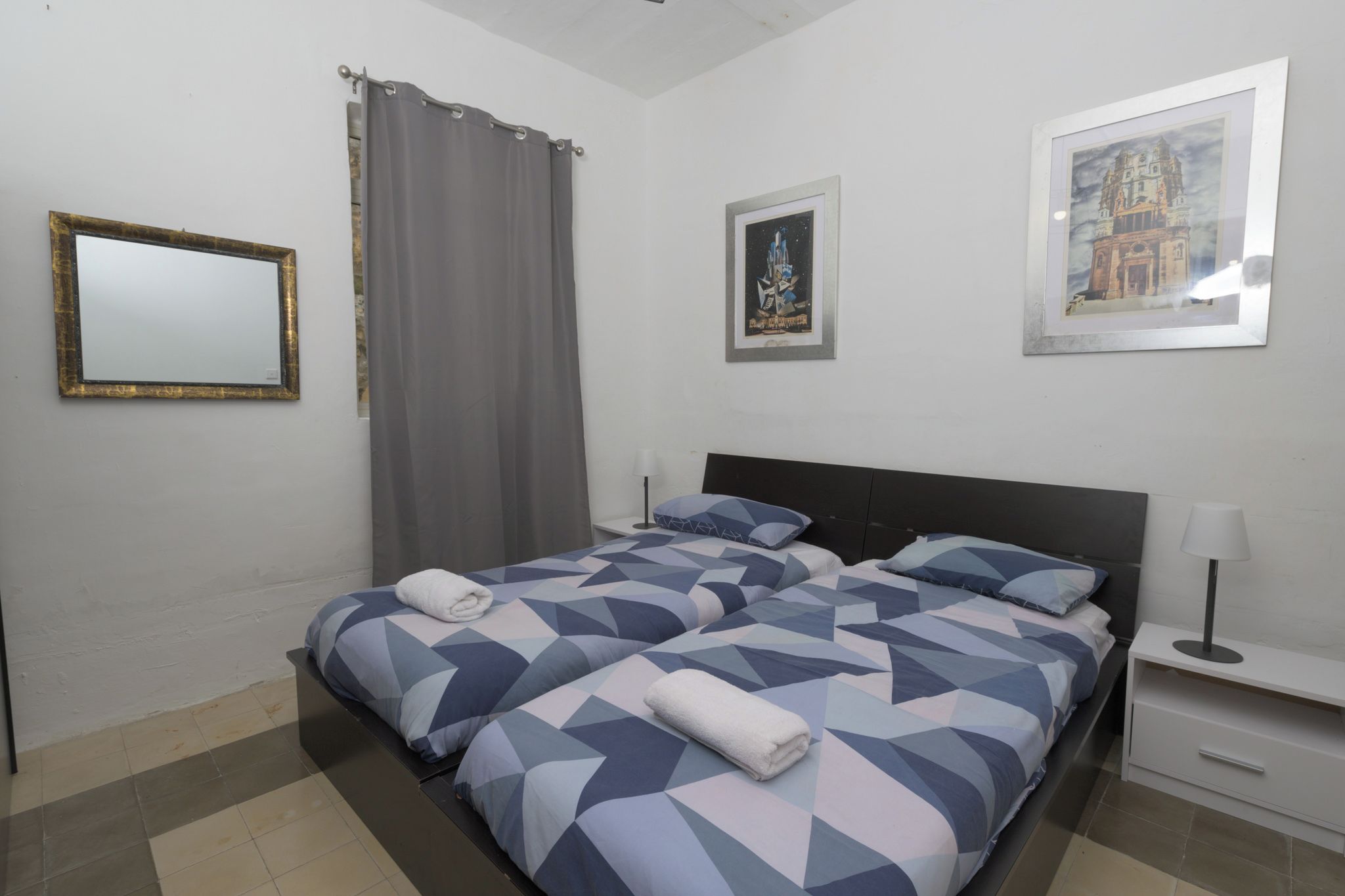A hotel room with a bed, a nightstand, a lamp, a mirror, and two framed pictures on the wall.