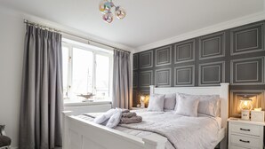 A bedroom with a white bed, grey curtains, a window, a bedside table with a lamp, and a wall with paneling. - Tritons Retreat (Moelfre)