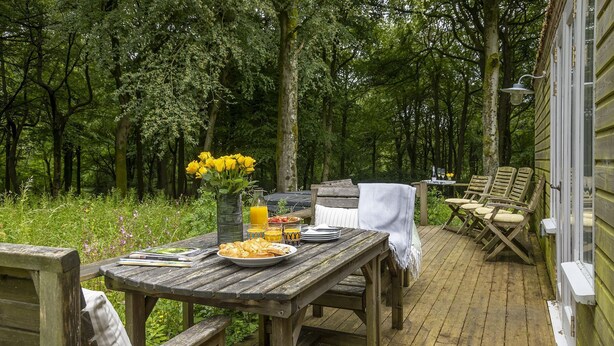 An outdoor wooden deck with a table set for breakfast, surrounded by chairs, and a bench. - Dog friendly holiday home with a hot tub and woodland views - Keepers Stable (Cranham)