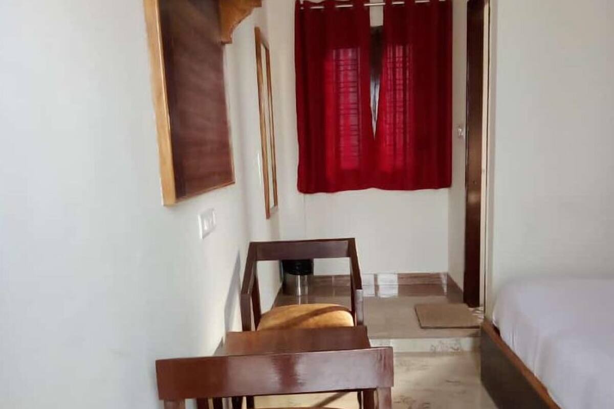 Ein schmaler Raum mit einem Bett, einem Holzstuhl, einem kleinen Tisch und einem roten Vorhang.