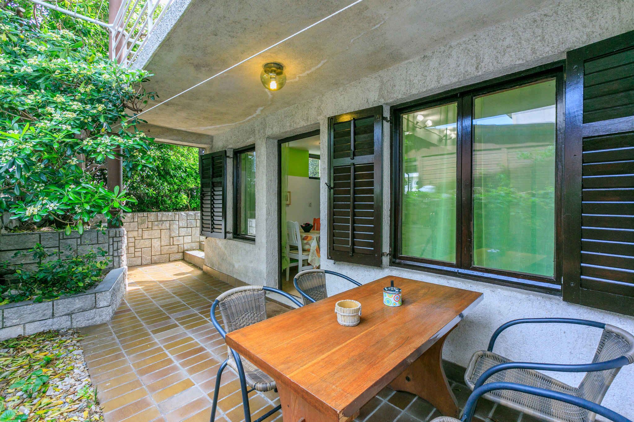 A patio area with a wooden table and chairs, a stone planter with greenery, and large windows with shutters.