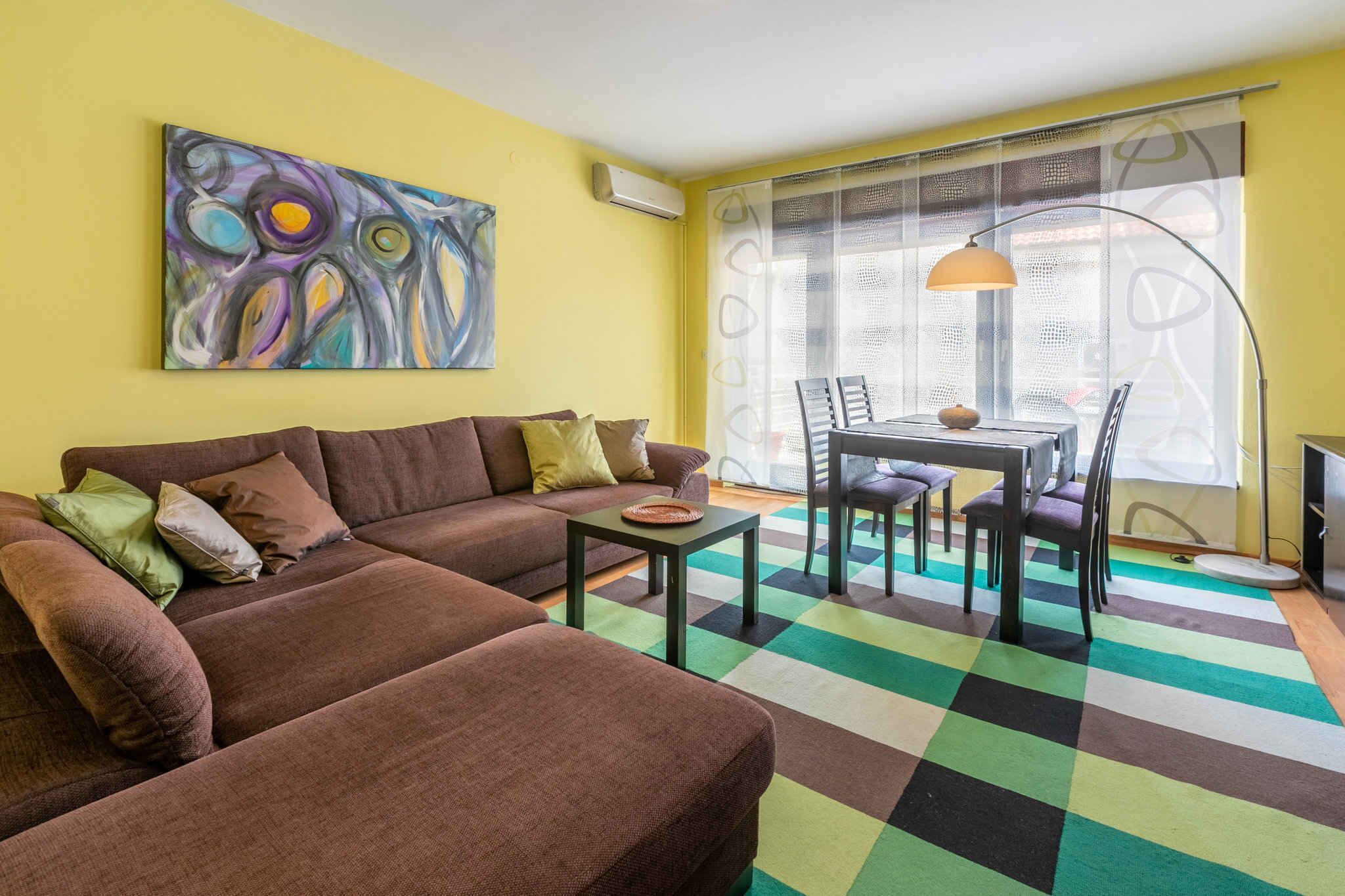A living room with a brown sofa, a dining table with chairs, a colorful rug, and a large abstract painting on the wall.