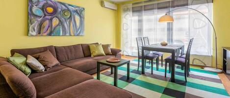 A living room with a brown sofa, a dining table with chairs, a colorful rug, and a large abstract painting on the wall.