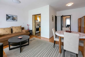 Ein Wohnzimmer mit einer Couch, einem runden Couchtisch und einem Esstisch mit Stühlen. Durch einen offenen Durchgang ist eine Küche erkennbar.