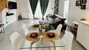 A modern living room with a glass dining table set for four, surrounded by white chairs. A staircase leads to an upper level, and a sofa is visible in the background.
