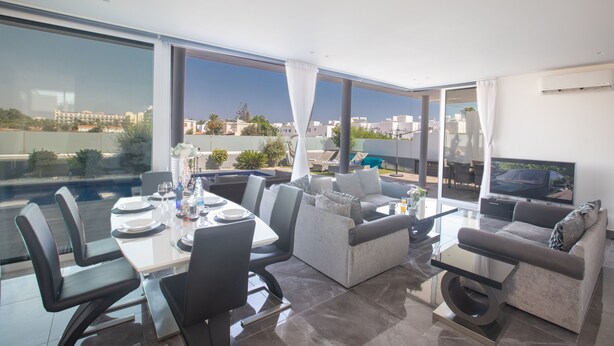 A modern living room with a dining area, large windows, and a flat-screen TV. - Villa Aria, Luxury 3BDR Protaras Villa with Private Pool, Close to the Beach (Protaras)