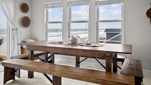 Et strandhus indvendigt med et stort træbord, bænk og udsigt over havet gennem store vinduer.