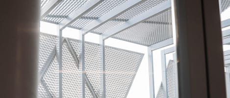 A modern architectural structure with a metal framework and patterned facade, viewed from an interior space with a wooden floor and a metal railing.