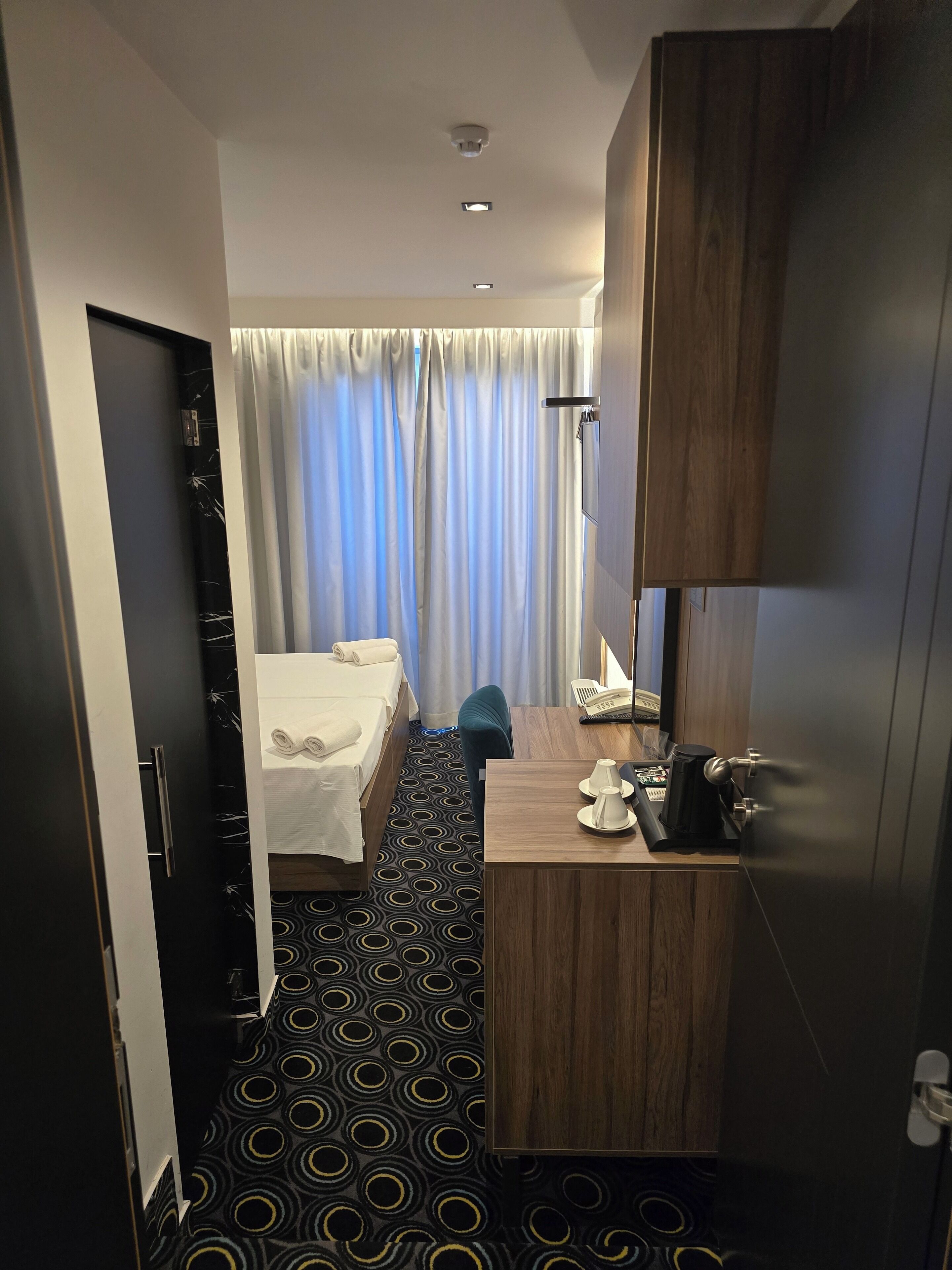 Quarto de hotel com cama, escrivaninha com telefone e cafeteira, banheiro com cortina de chuveiro e piso estampado.
