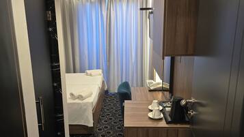 Quarto de hotel com cama, escrivaninha com telefone e cafeteira, banheiro com cortina de chuveiro e piso estampado.