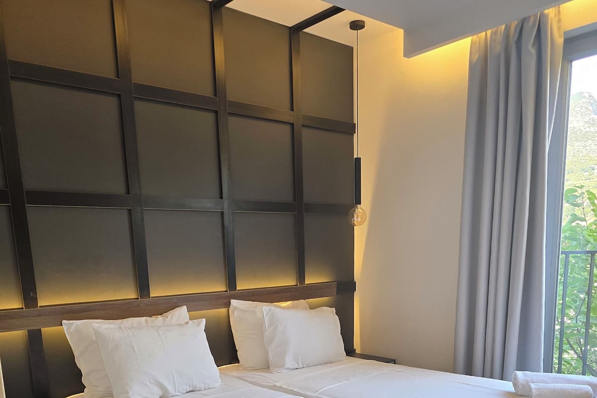 A hotel room with a large bed, two pillows, a bedside table, a lamp, and a window with curtains.