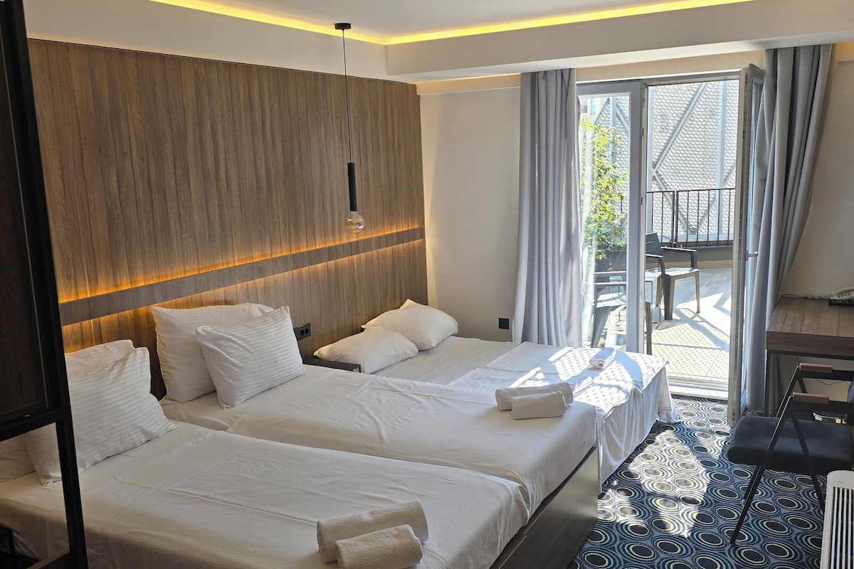 A hotel room with two beds, a wooden headboard, a large window with curtains, a patterned floor, and a small balcony area.