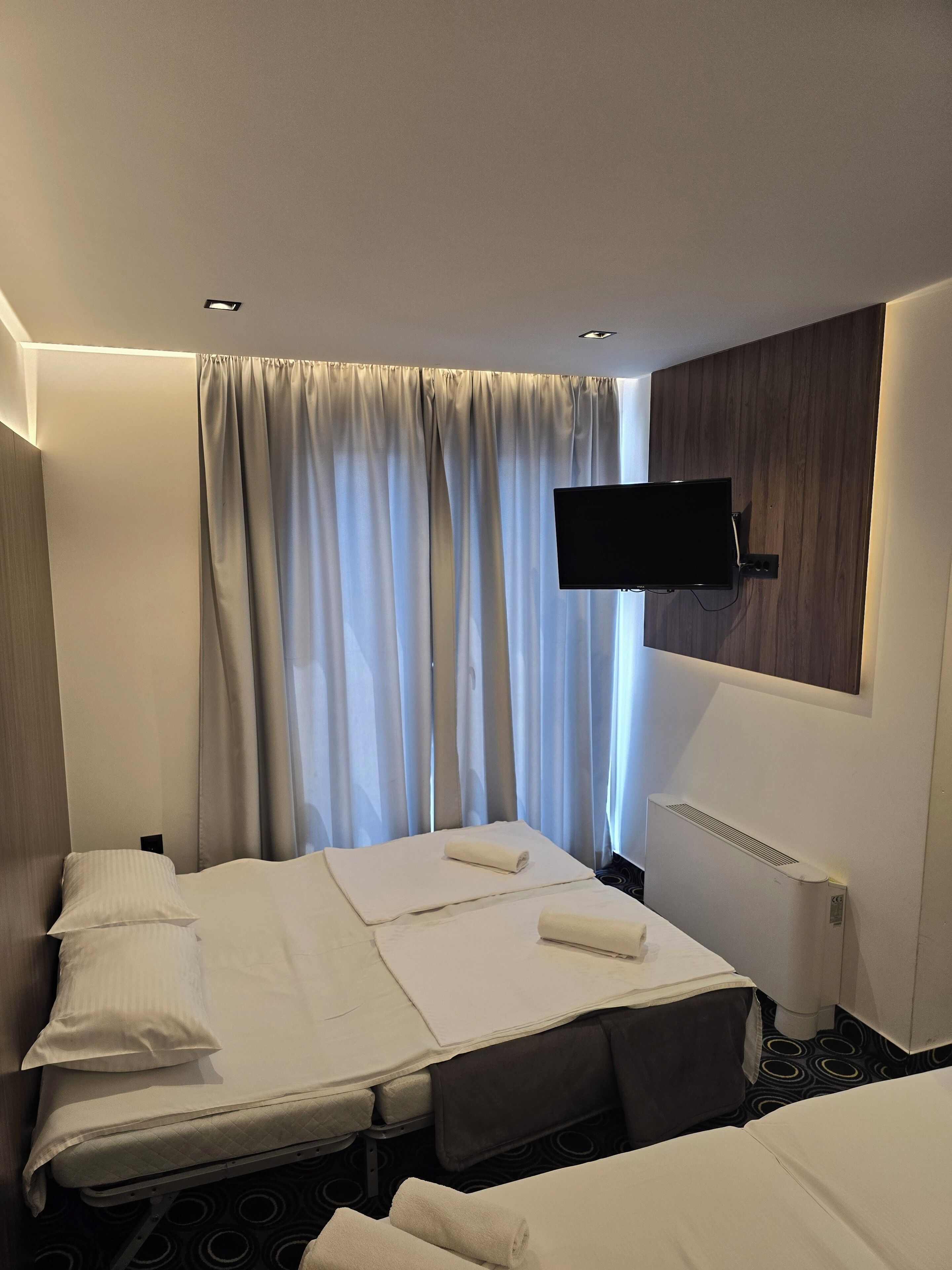 Habitación de hotel con cama, televisor de pantalla plana montado en la pared, ventana con cortinas y radiador.