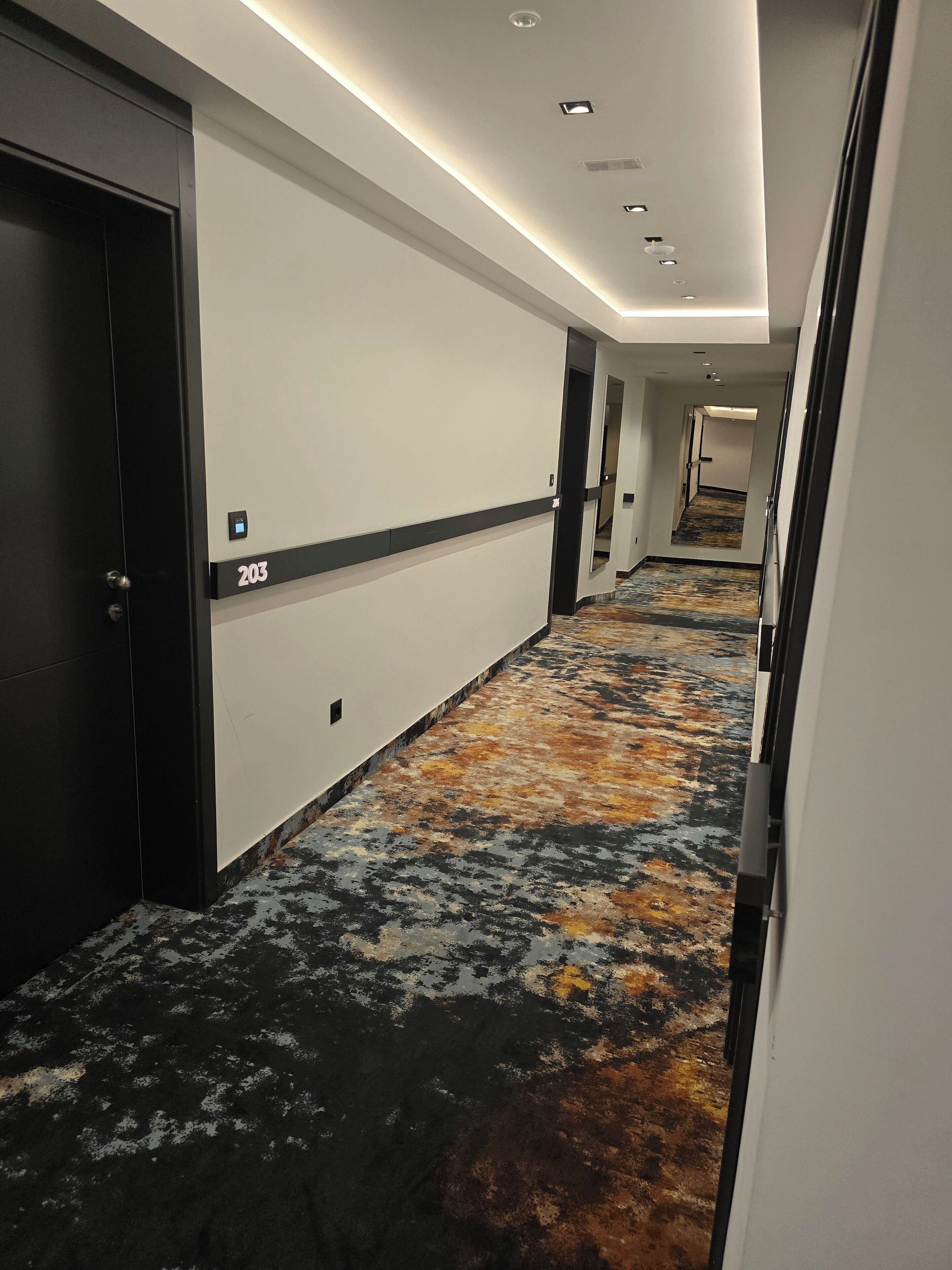 Ein Hotelkorridor mit Teppichboden, einer Tür mit der Bezeichnung '203' und einem Gang, der zu einem anderen Zimmer führt.