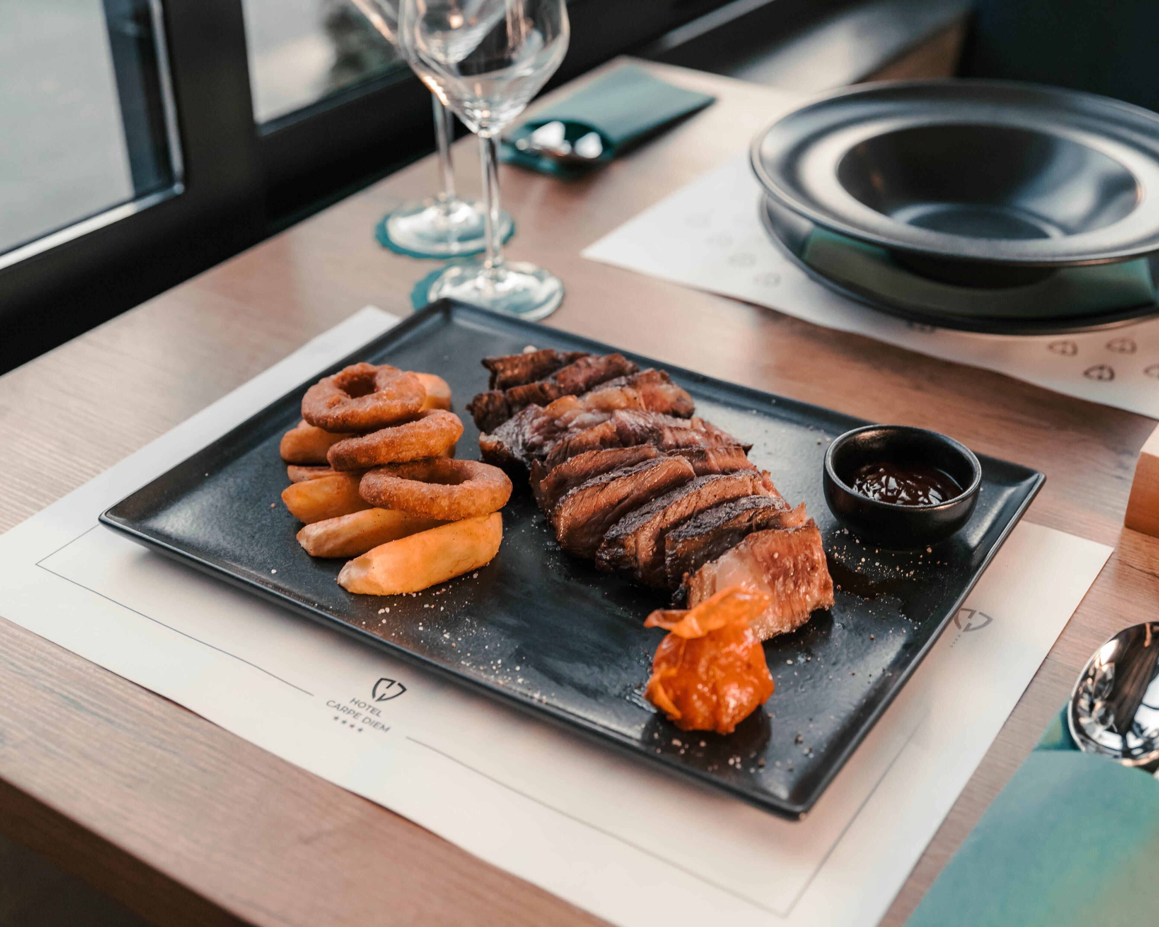 Une assiette noire contenant un plat composé d’un steak, de rondelles d’oignon et d’une petite portion de sauce, posée sur une table. On aperçoit en arrière-plan un menu et un verre d’eau.